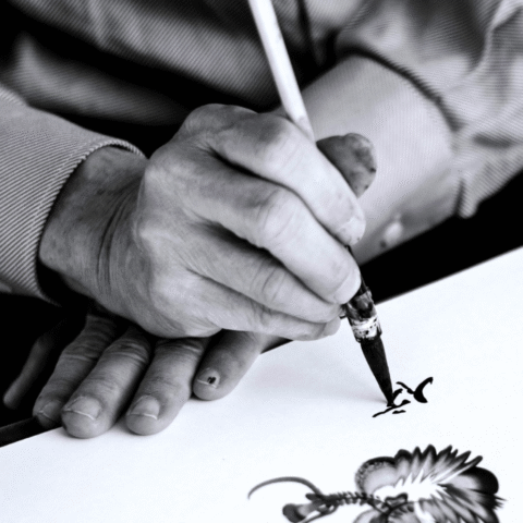 A black and white photo of someone's hands working on a Chinese drawing and caligraphy.