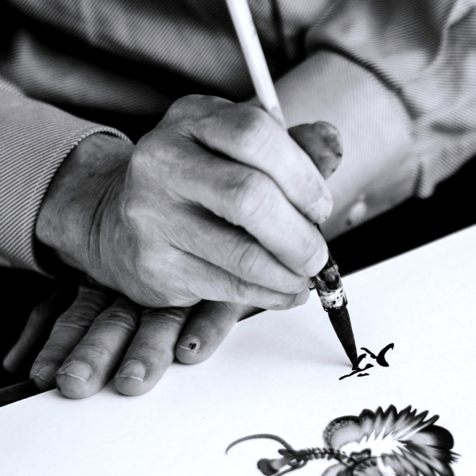 A black and white photo of someone's hands working on a Chinese drawing and caligraphy.