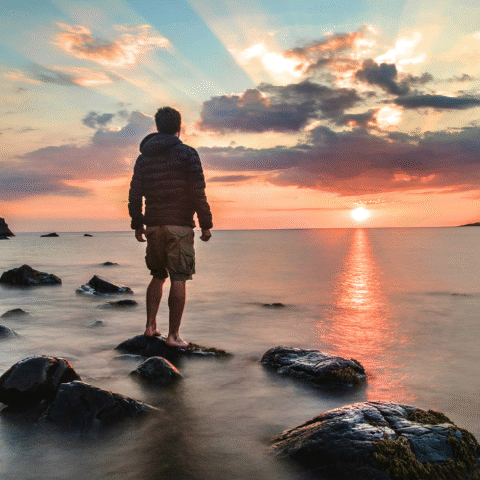 A man standing on a rock near the shore of the ocean looking at the sun set.