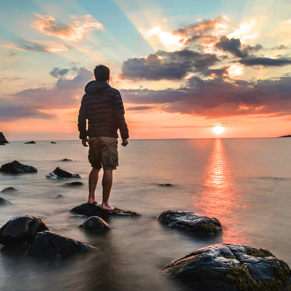 A man standing on a rock near the shore of the ocean looking at the sun set.